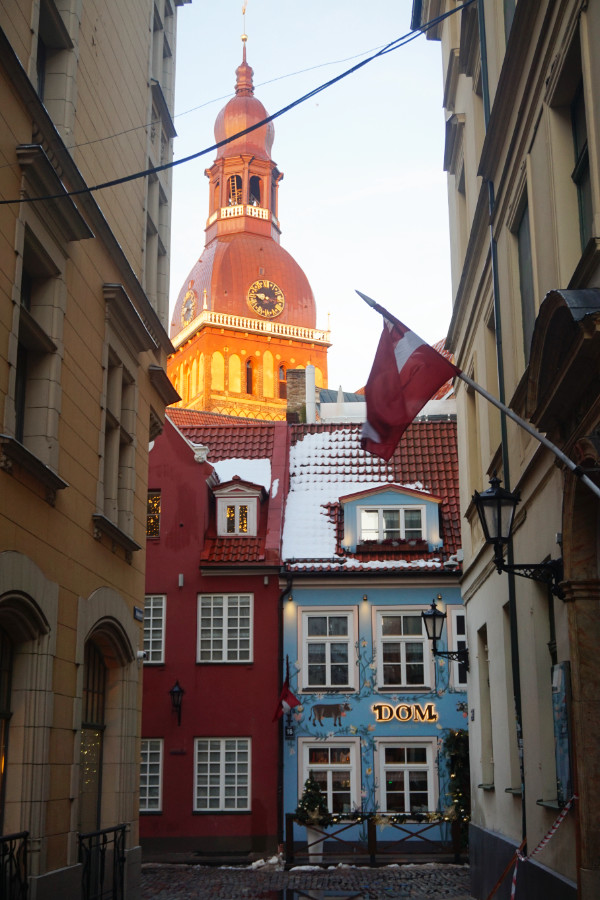 Alleyway in Riga, Latvia, with the Dom spire in the background behind two colourful buildings (red and blue), and a Latvian flag hanging to the right hand side.