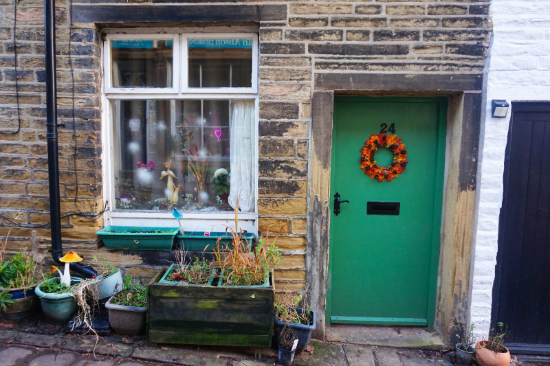 House in Haworth, Yorkshire. To the right is a green door with an autumn style wreath. On the left is a window with lights and some planters in front.